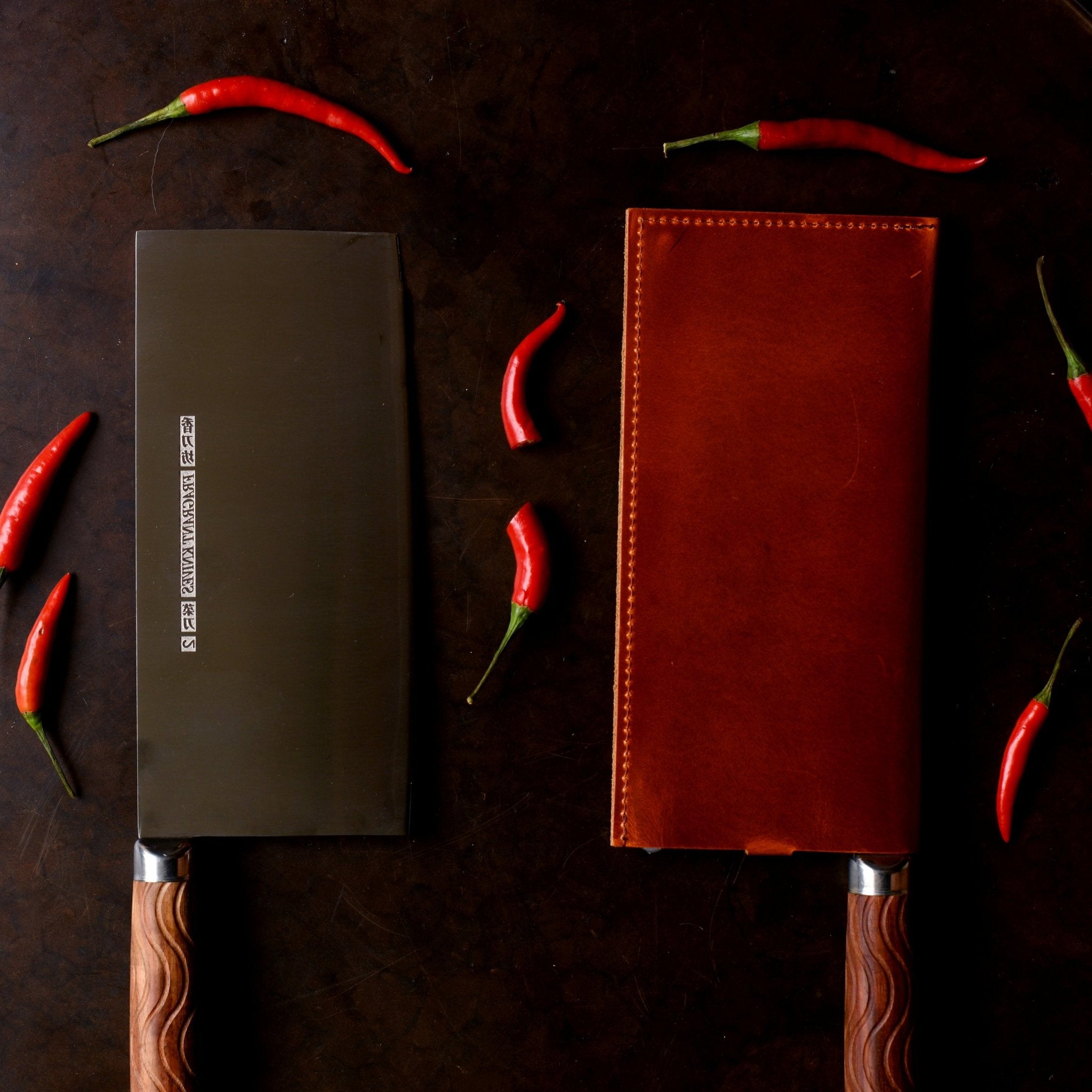 Two knives, one in brown leather sheath, on a dark surface with red chilies.
