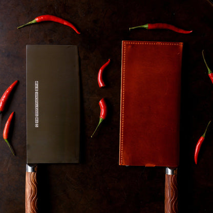 Two knives, one in brown leather sheath, on a dark surface with red chilies.